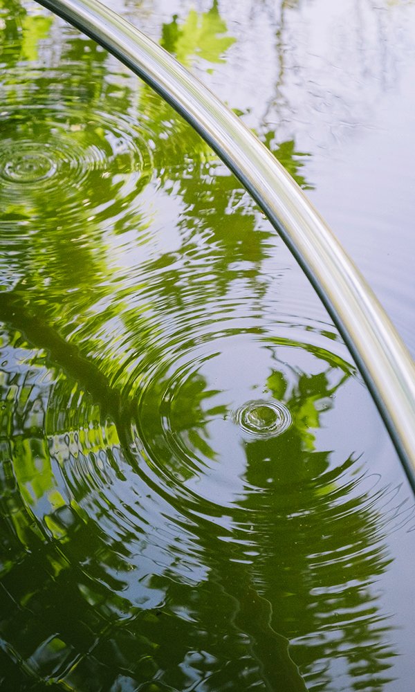 Une goutte touche l'eau au pied du tube en inox, reflets verts des arbres